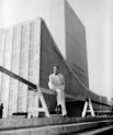 A man posing and sitting in  front of a large building with glass windows (UN headquarters)