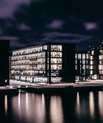 Dark photo, evening. Big buildings placed close to the harbour. Windows from the buildings light up.