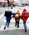 three children running across the street wearing school backpacks.