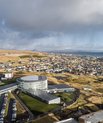 Picture taken from up high, propably a helicopter. Shows the city of Tórshavn and Glasir. The Glasir building is round shaped with glass windows all around. Three rectangular buildings are then connected all around the building.