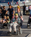 People of all ages walking on a bridge together. Two elderly people on wheelchairs.